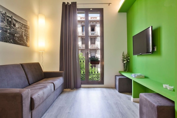 A modern living room with a sofa, a TV mounted on a green wall, a window with a view of a balcony, and a door.