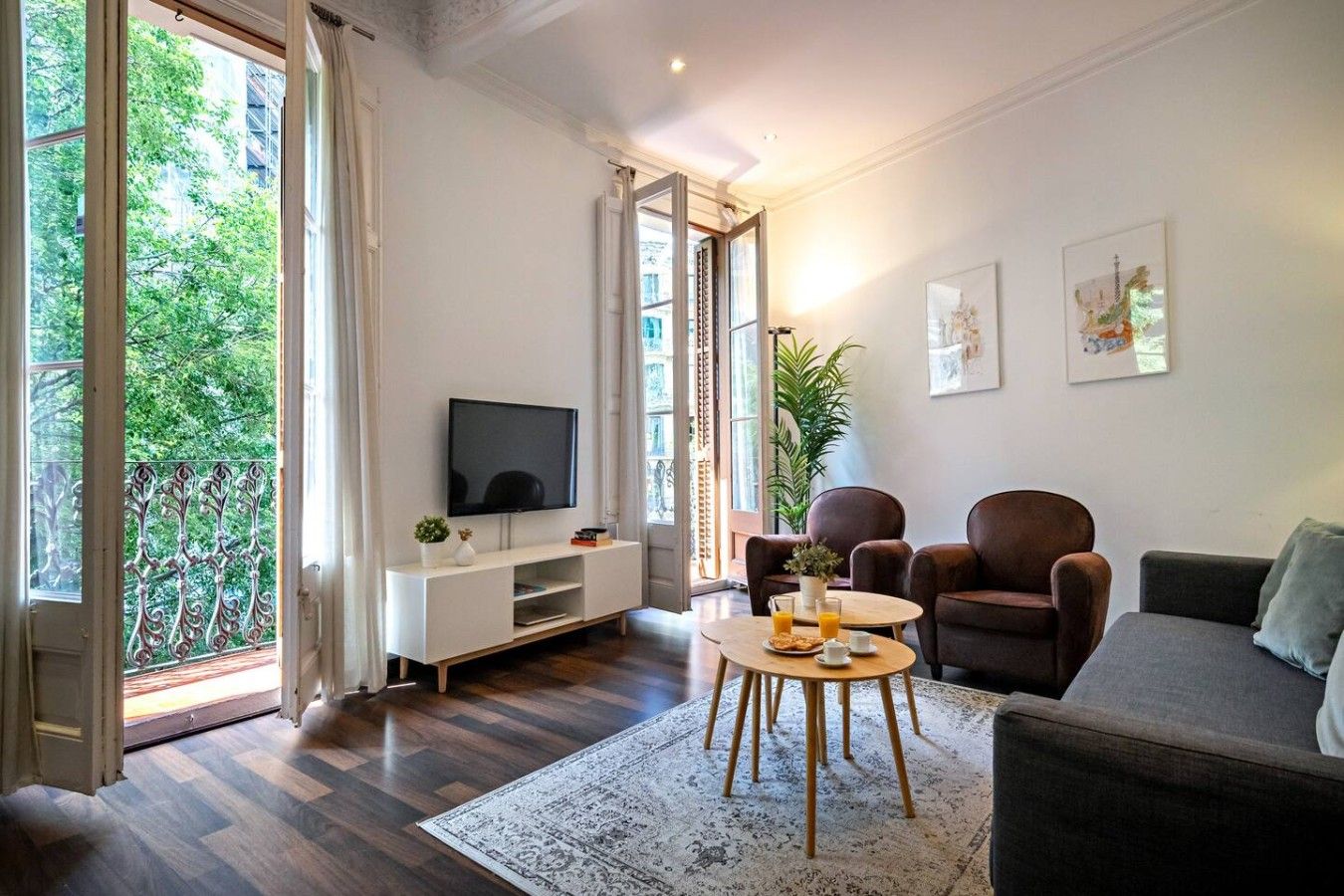 A living room with a flat-screen TV, a sofa, two armchairs, a coffee table, and a dining table set for breakfast.