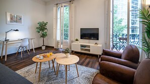 A modern living room with a sofa, a TV, a dining table with chairs, a desk with a chair, and a potted plant.