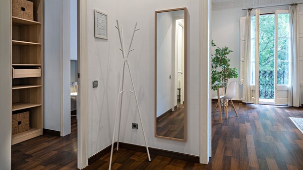 A hallway with wooden flooring, a white wall-mounted mirror, and a door leading to a room with a large window.