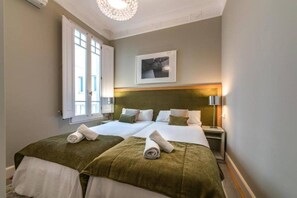 A hotel room with two beds, a large window, a chandelier, and a framed picture on the wall.