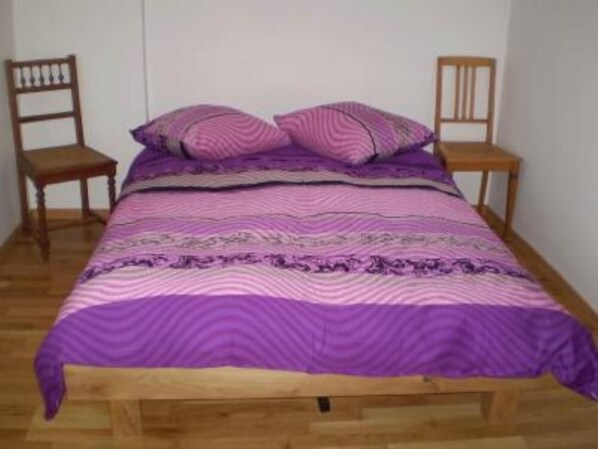 Una cama con colcha estampada en tonos morado y rosa, flanqueada por dos sillas de madera.