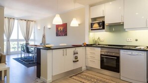 A modern kitchen with white cabinets, a built-in oven, and a dishwasher. There is a dining area with a table and chairs, and a large window with curtains. - Cascais Panoramic with Pool by Portugal Portfolio (Cascais)