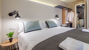 Una habitación de hotel moderna con una cama bien hecha, una mesita con una planta y una kitchenette al fondo.