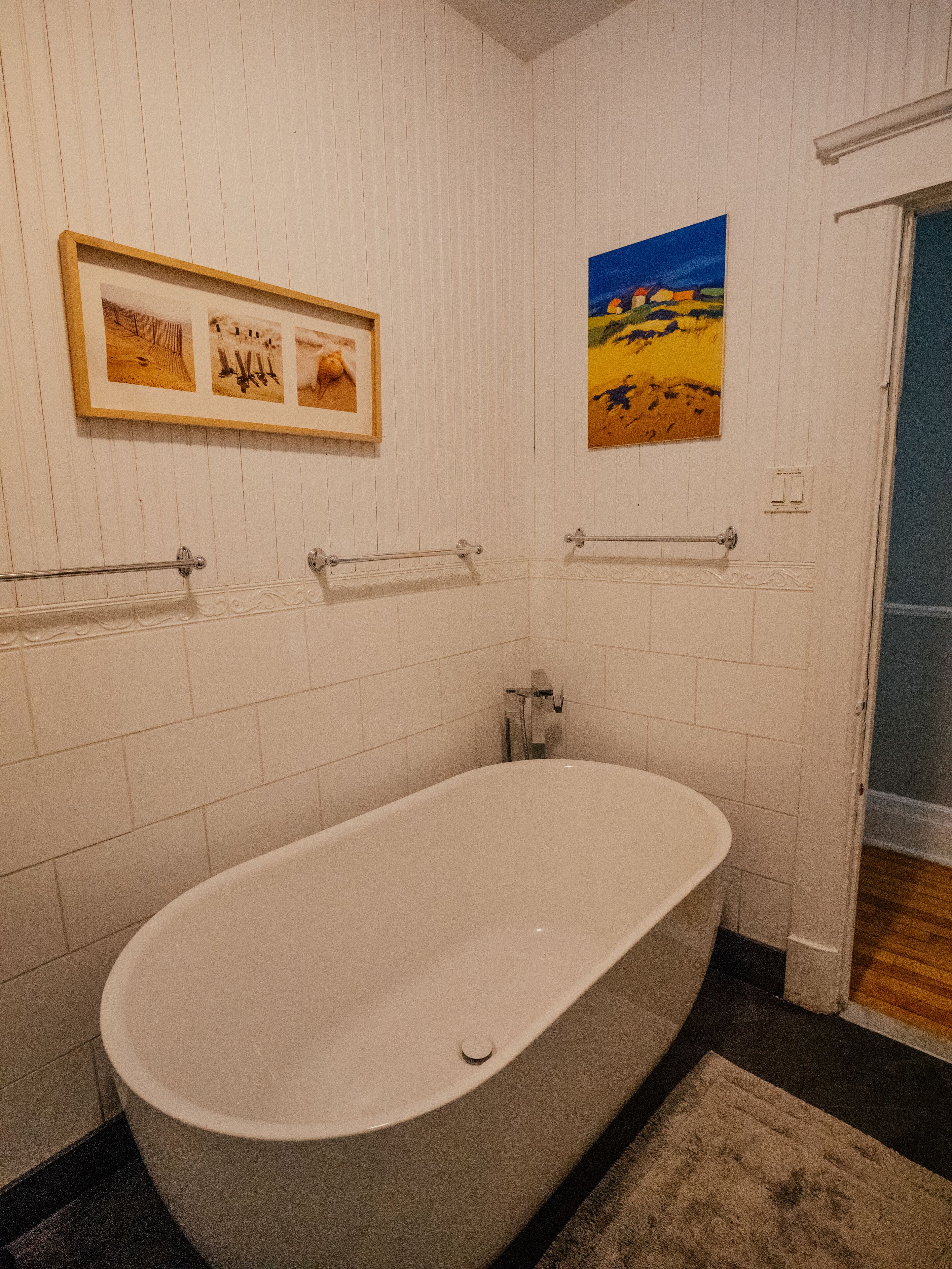 Une salle de bain avec une baignoire sur pieds, des murs recouverts de carreaux blancs, une photographie encadrée et un tableau représentant un paysage.