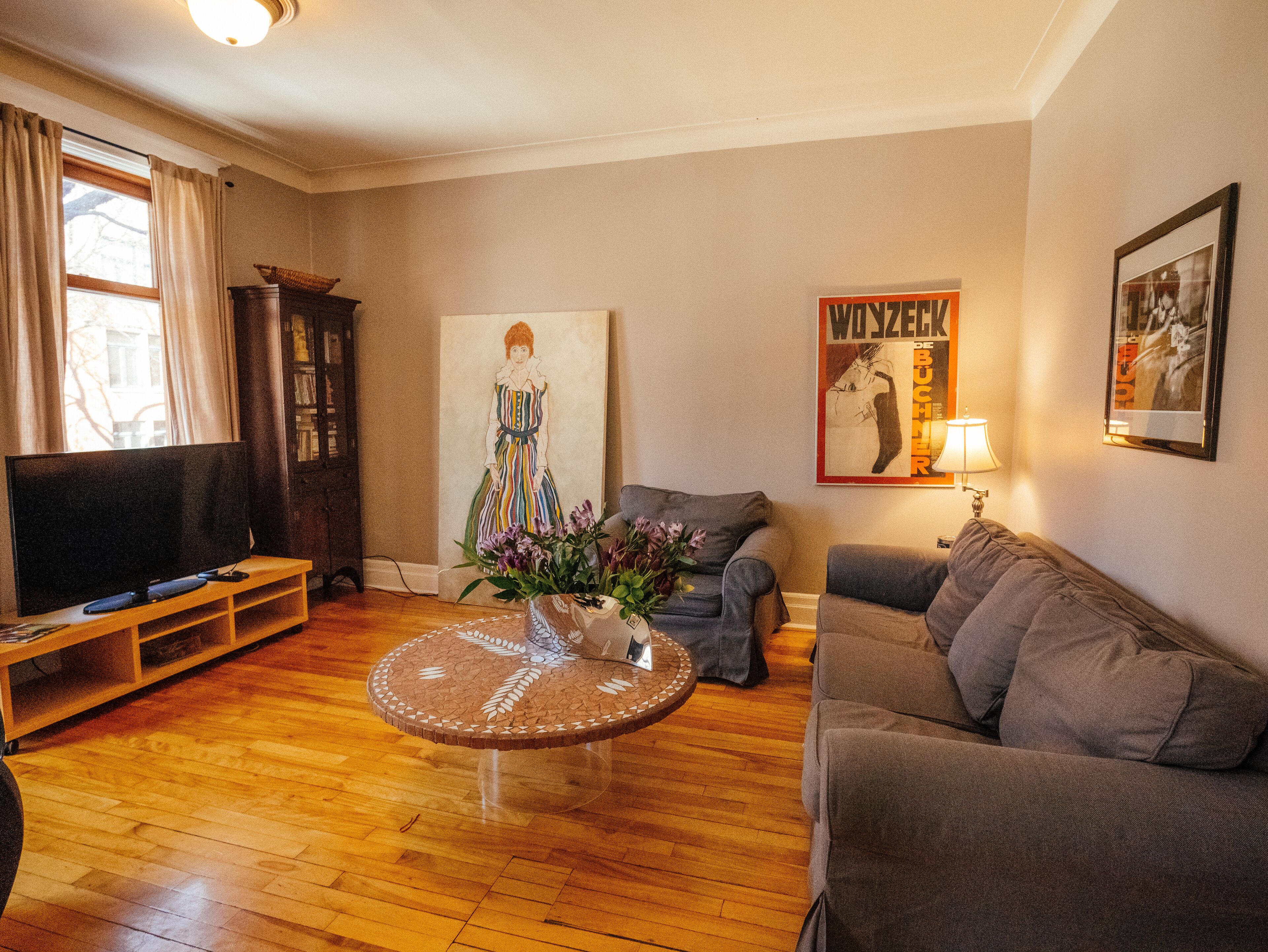 Una sala de estar con piso de madera, televisor, sofá, una mesa de centro con un jarrón de flores y un cuadro en la pared.