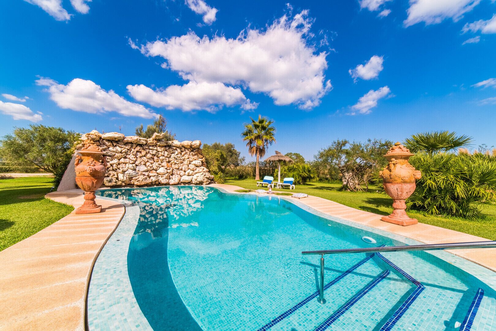 A swimming pool with a stone wall and two urns, surrounded by a well-maintained lawn and palm trees.