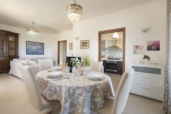 A dining area with a table set for four, a chandelier, and a kitchen in the background.