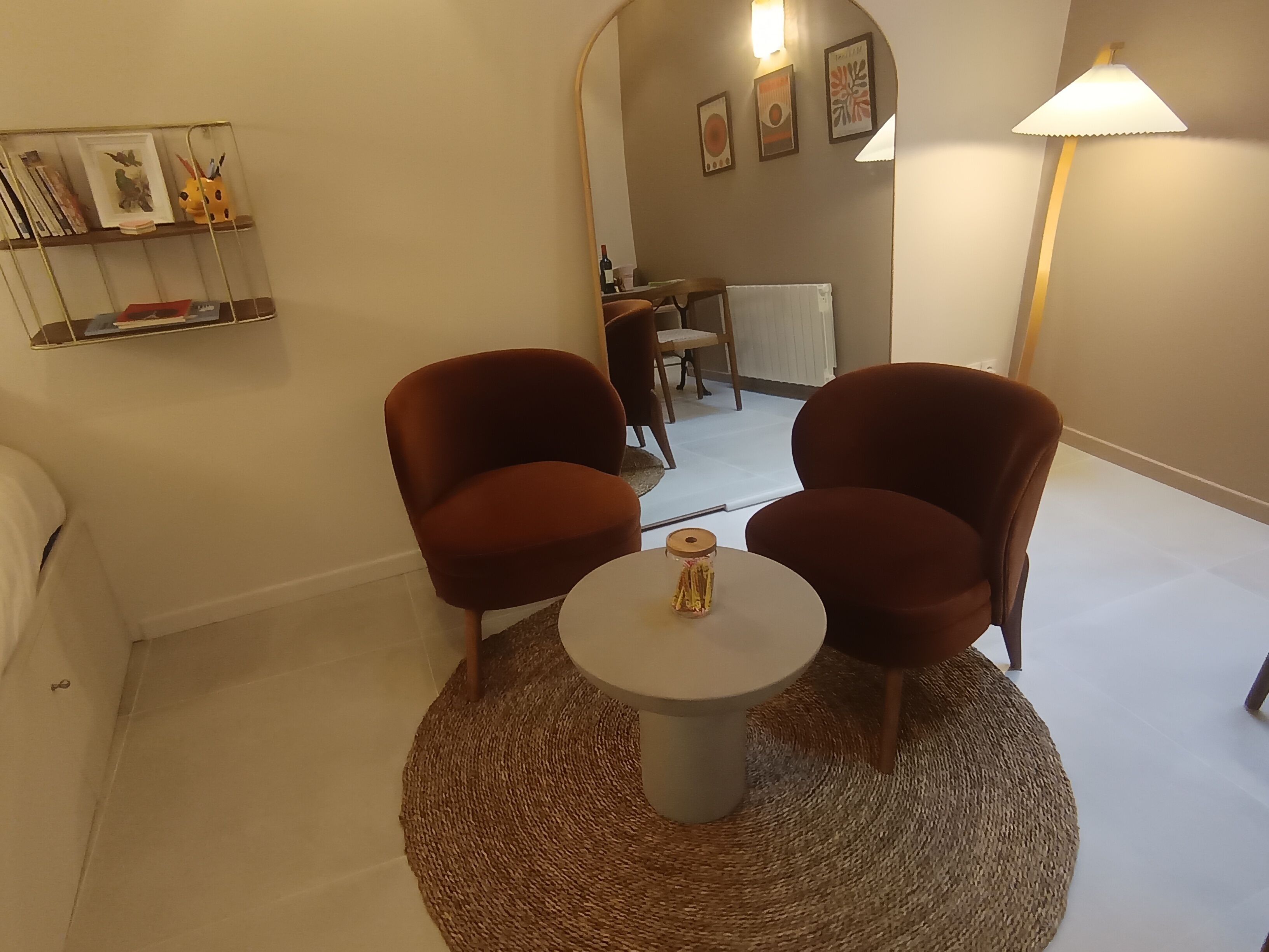 Een gezellige kamer met twee bruine fauteuils, een klein rond tafeltje en een lamp. Er is een plank met boeken en een lijst met een foto aan de muur.
