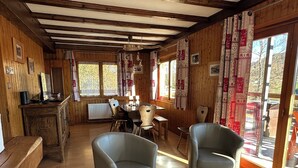 Ein gemütliches Blockhaus-Interieur mit Holzwänden, einem Essbereich mit Tisch und Stühlen sowie großen Fenstern mit Vorhängen.