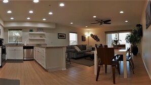 Eine moderne Küche mit weißen Schränken, einem Edelstahlkühlschrank und einem Geschirrspüler. Es gibt einen Essbereich mit Tisch und Stühlen, und ein graues Sofa im Wohnzimmer.