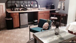Una cocina compacta con refrigerador, microondas y cafetera. Hay una silla color turquesa y una mesa de madera con un florero.