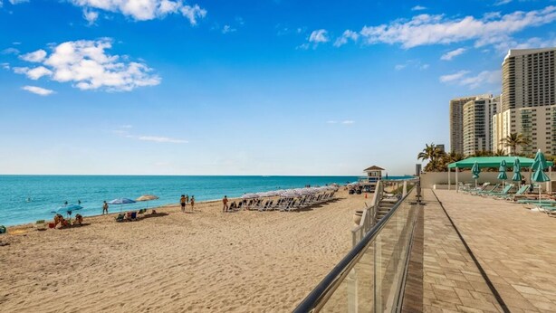Ein Strand mit blauem Himmel, sandigem Ufer und einer Reihe von Strandliegen und Sonnenschirmen.