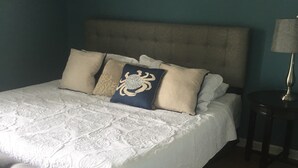Una cama bien hecha con una colcha blanca y varias almohadas, una mesita de noche con una lámpara y un adorno en la pared.