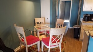 Un comedor con una mesa de vidrio rodeada por sillas de mimbre con cojines rojos, una alfombra estampada y una zona de cocina con cafetera y cubo de basura.