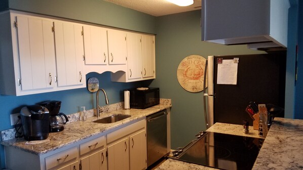 Cocina compacta con armarios blancos, microondas negro, lavavajillas de acero inoxidable y refrigerador con una nota adherida.
