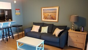 Una sala de estar con un sofá azul, una mesa de centro blanca, una mesita de noche de mimbre y un cuadro de una playa en la pared.
