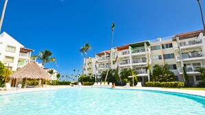 A resort pool area with a clear blue sky, palm trees, and a thatched-roof hut. - Oceanfront 2Bd/2Ba Penthouse Condo  (Bavaro)