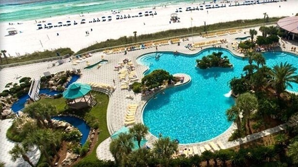 A large, irregularly shaped swimming pool surrounded by palm trees and lounge areas, with a beach and ocean in the background. - 'Osprey's Nest'  Lower prices for August! Lovely condo, close to attractions. (Panama City Beach)