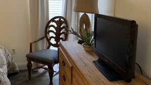 A room with a wooden dresser, a television on top, a chair, a potted plant, and a window with curtains. - 'Osprey's Nest'  Lower prices for August! Lovely condo, close to attractions. (Panama City Beach)