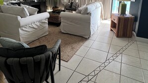 A living room with two white sofas, a black armchair, a television, a large plant, and a view of the outdoors through a window. - 'Osprey's Nest'  Lower prices for August! Lovely condo, close to attractions. (Panama City Beach)
