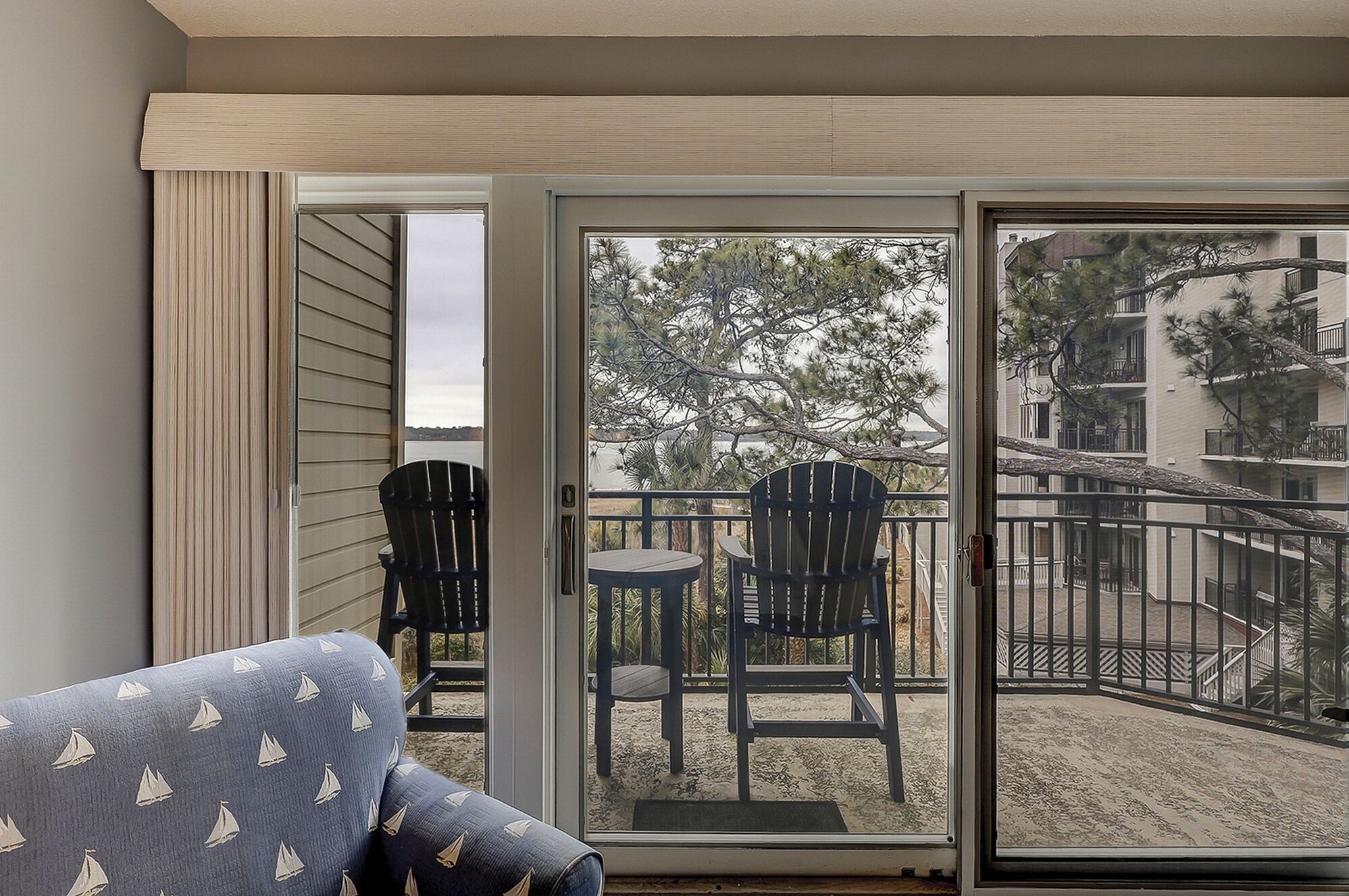 A balcony with a sliding glass door, a small round table, two Adirondack chairs, and a view of a building and trees.
