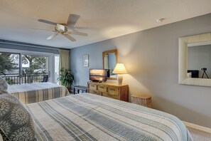 Un dormitorio con cama, ventilador de techo, televisor, lámpara, espejo y ventana con cortinas.