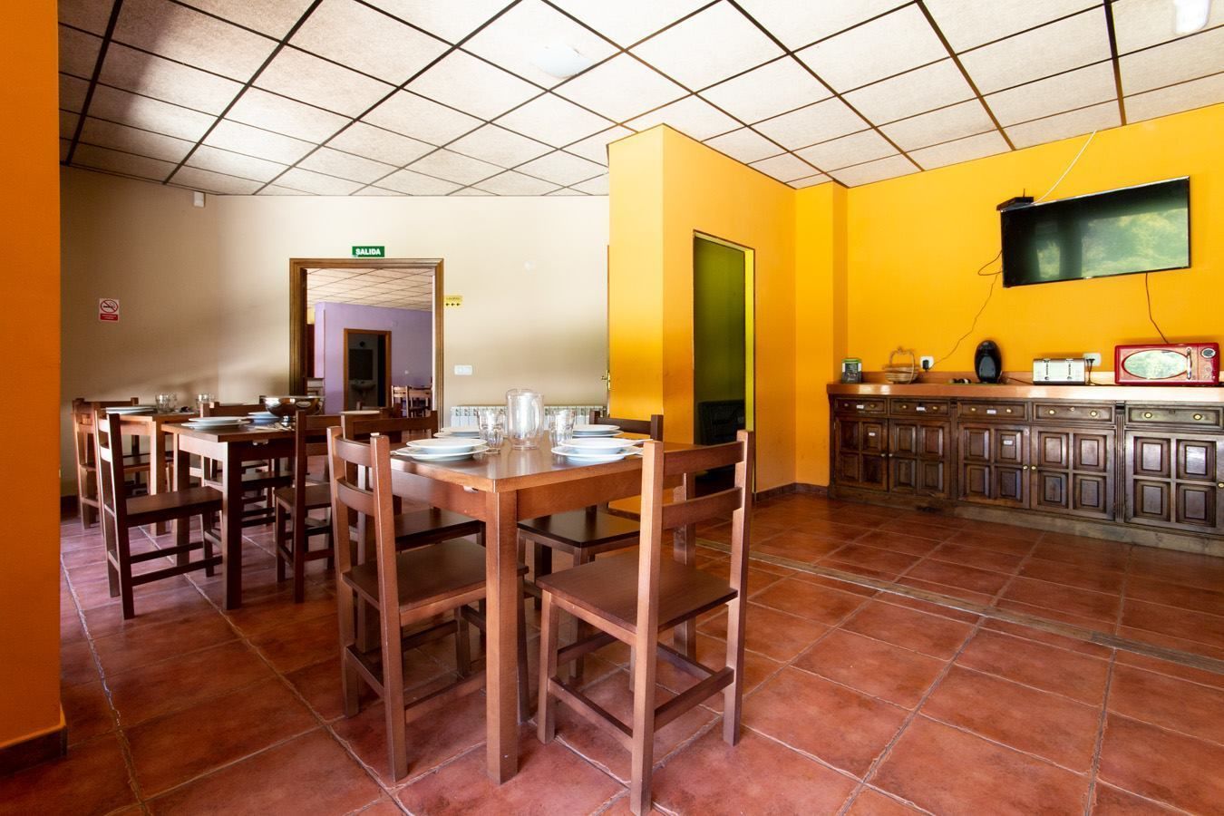A dining area with wooden tables and chairs, a tiled floor, a yellow wall, and a television mounted on the wall.
