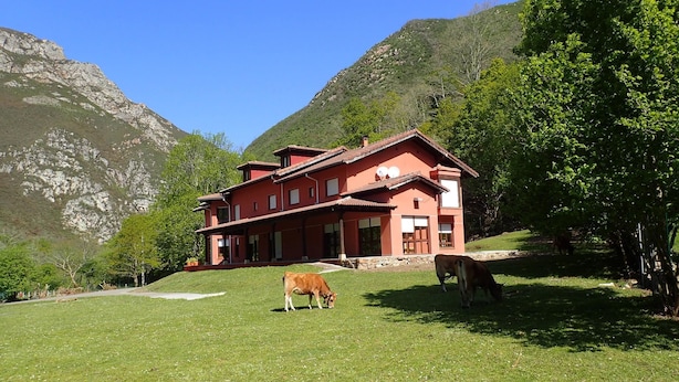 A red building with a sloped roof, surrounded by a grassy area and trees, with mountains in the background. - Albergue Los Avellanos - Hostel (Pilona)