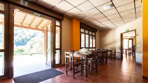 A restaurant with a tiled floor, wooden tables and chairs, and a view of the outdoors through large windows. - Albergue Los Avellanos - Hostel (Pilona)