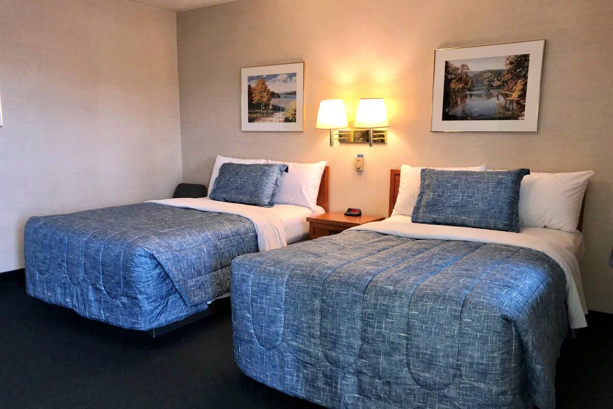 A hotel room with two beds, each with a blue quilted bedspread, white pillows, and a nightstand with a lamp between the beds.