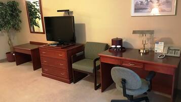 A hotel room with a wooden desk, a television, a green chair, a desk lamp, a telephone, and a framed picture on the wall.