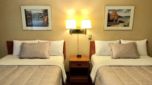 A hotel room with two beds, each with a pillow and a lamp between them. There are framed pictures on the wall above the beds.