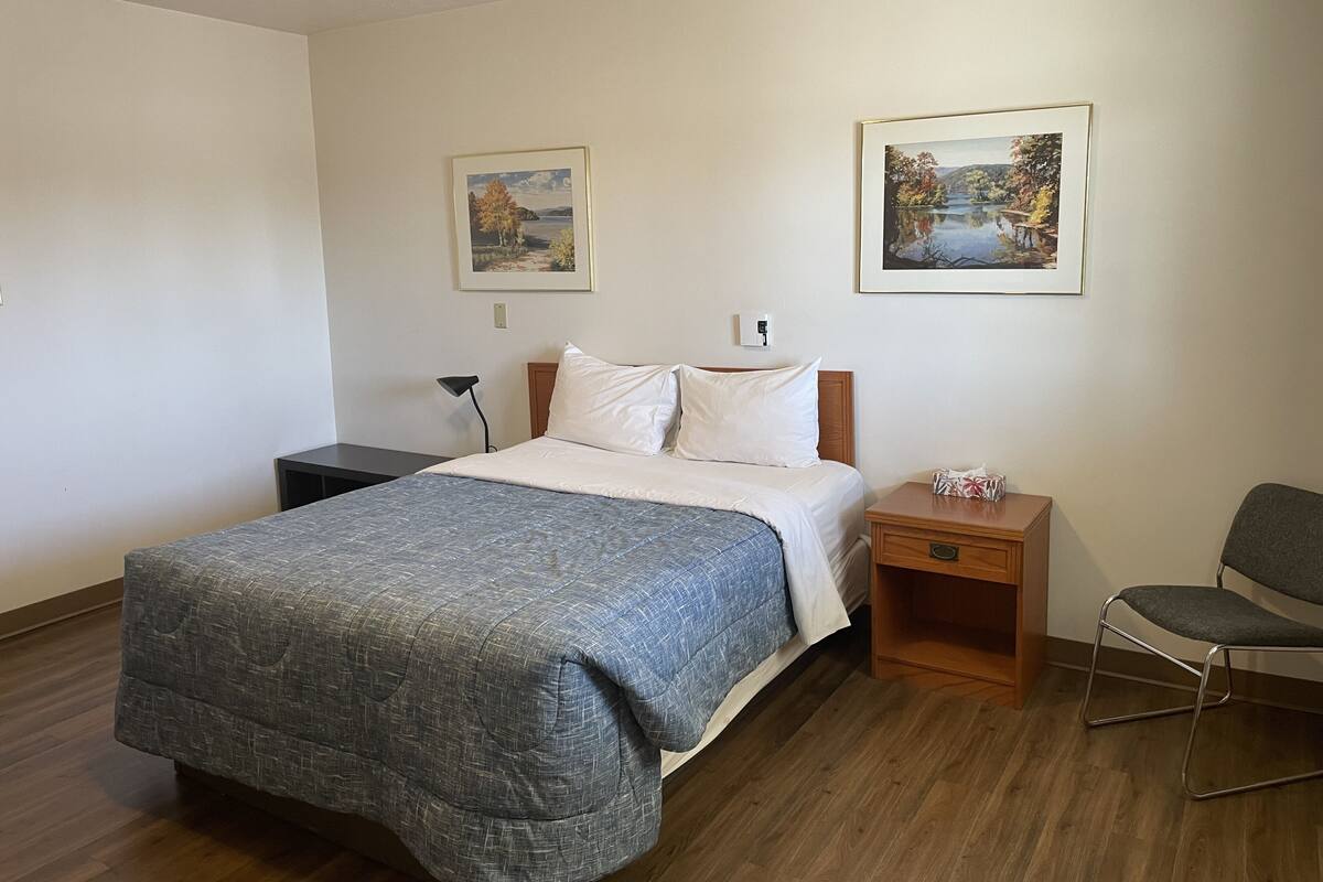 A neatly arranged hotel room with a bed, a bedside table, a chair, and two framed pictures on the wall.
