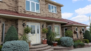 A two-story house with a brick facade, a red roof, and a front porch with steps. There are potted plants and shrubs in front of the house.