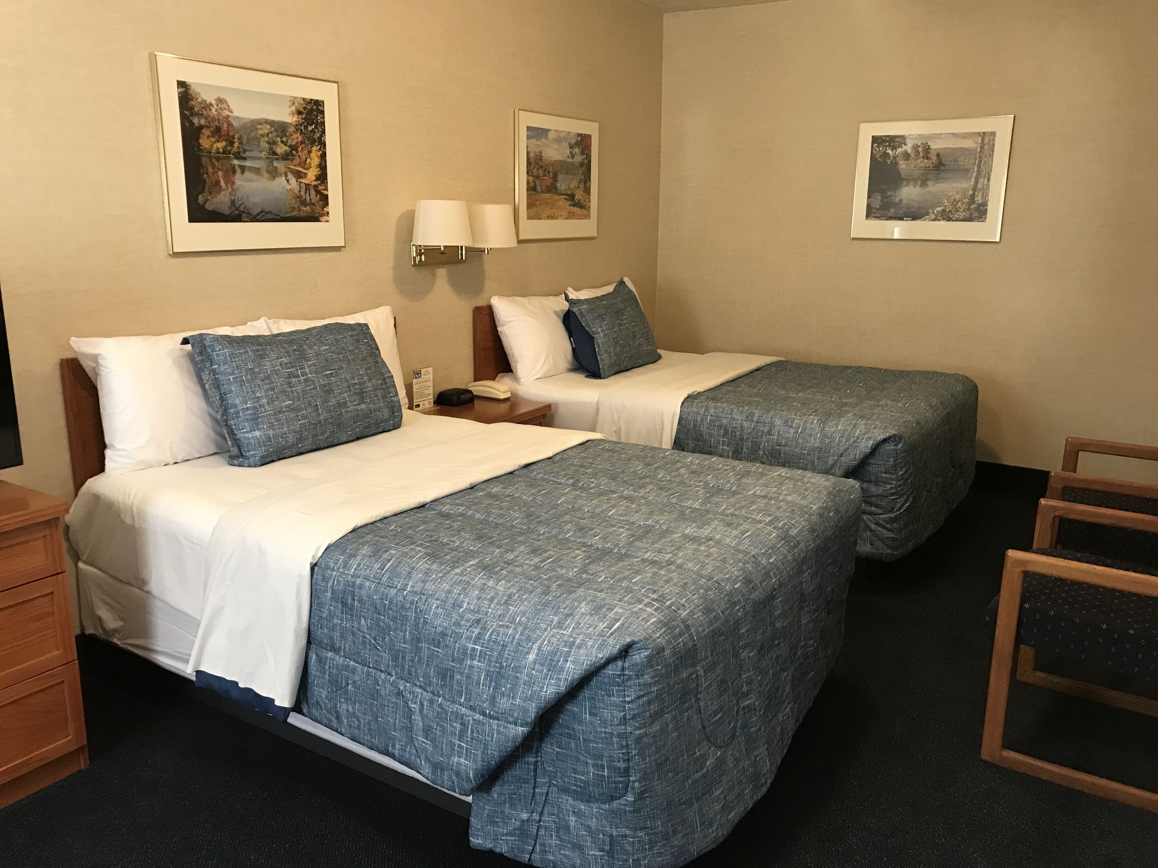 A hotel room with two beds, a nightstand, a lamp, and framed pictures on the wall.