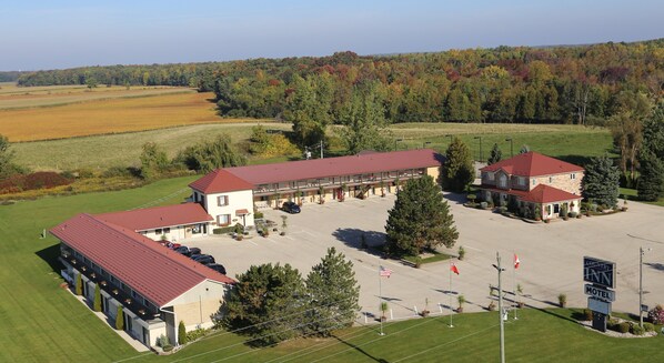 A hotel with a central building and two wings, a parking lot, and surrounding fields and trees.