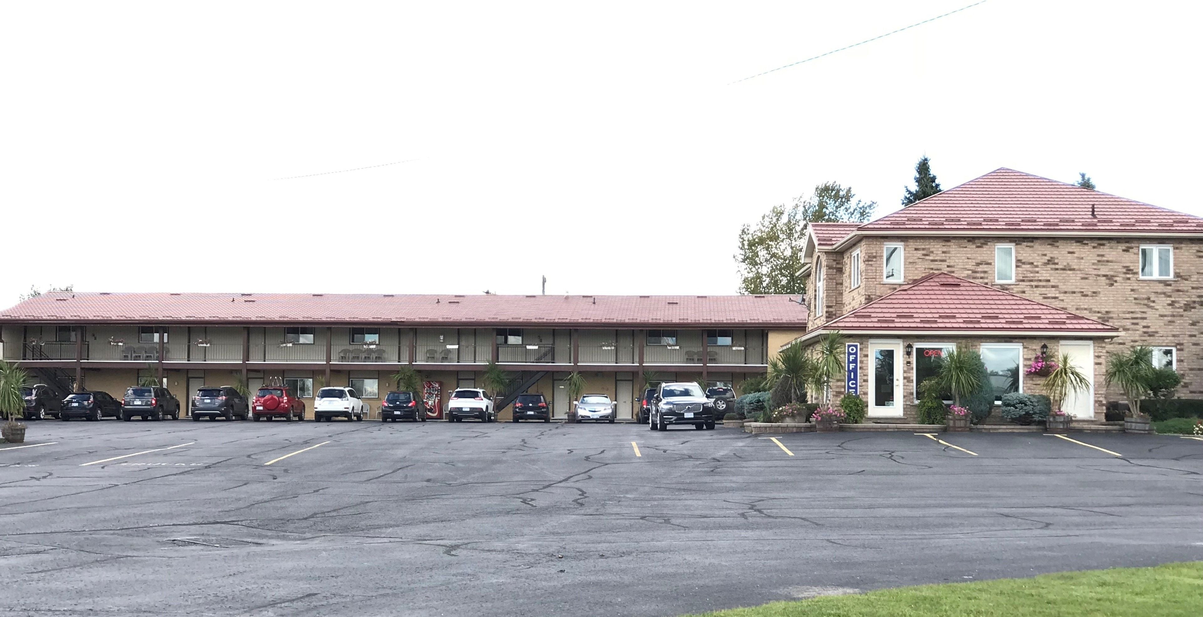 Een parkeerplaats met meerdere auto's, een motelgebouw met verschillende kamers, en een losstaand kantoorgebouw met een rood dak.