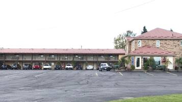 Een parkeerplaats met meerdere auto's, een motelgebouw met verschillende kamers, en een losstaand kantoorgebouw met een rood dak.
