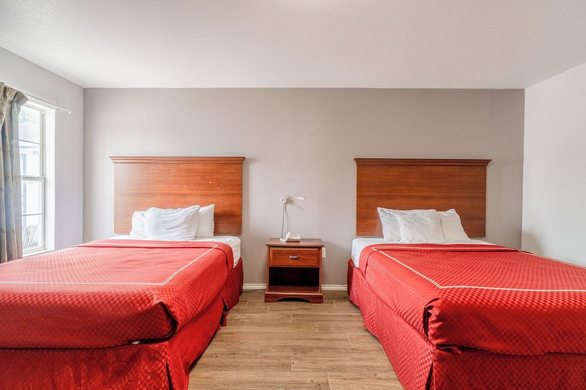 A hotel room with two single beds, wooden headboards, a nightstand with a lamp, and a window with curtains.