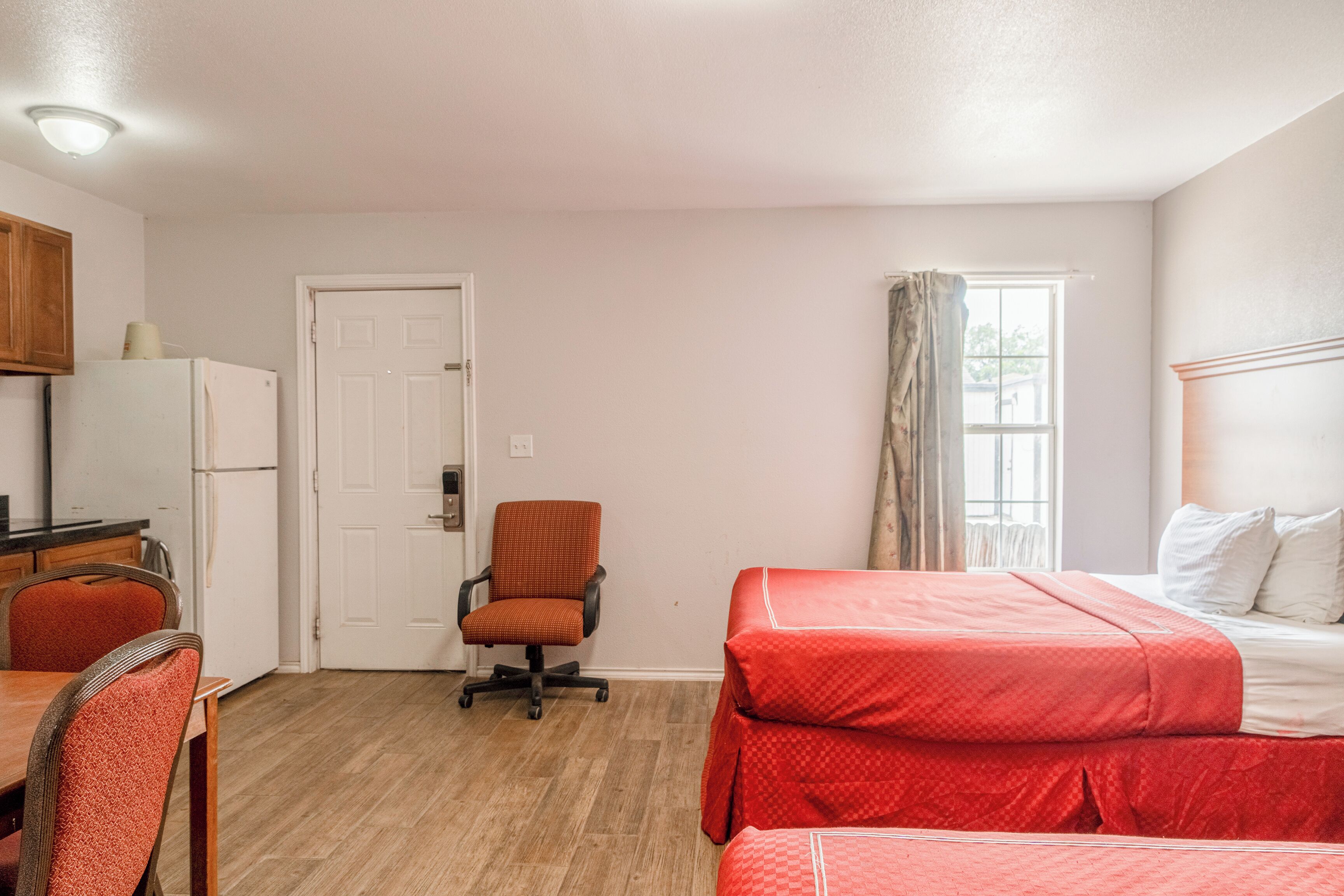 Habitación pequeña con kitchenette, una cama individual con ropa de cama roja, una mesa de comedor de madera con sillas y una ventana con cortinas.