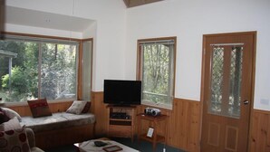 A cozy living room with a sofa, a television, and a small table. There are large windows with blinds, and a wooden door with a window.