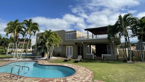 A pool area with a stone border, palm trees, and a two-story building with balconies. - Luquillo Beach- Puerto Rico, 4 Bedroom Vacation Villa With Pool and Wifi (Luquillo)