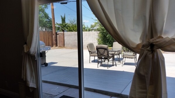 Blick durch eine Glastür mit Vorhängen auf eine Terrasse im Freien mit Tisch und Stühlen, einem Grill und einem Holzzaun.