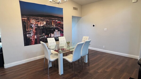 A dining area with a glass table and four chairs, a cityscape painting on the wall, and a chandelier. - STATE/CITY LICENSED AND PERMITTED! 3 bed 2 bath Single Family Home Pet Friendly. (Las Vegas)