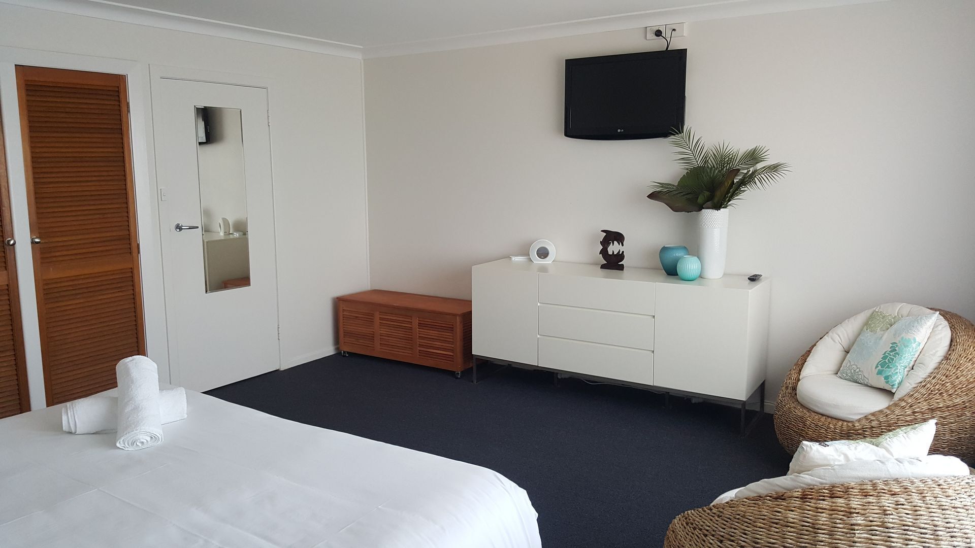 Una habitación de hotel con una cama, un banco de madera, un aparador blanco, un televisor y una silla de mimbre.