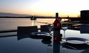 Een tafel voor twee, gedekt met een fles wijn en twee glazen, met uitzicht op een wateroppervlak bij zonsondergang.