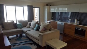 Una sala de estar moderna con un sofá beige, una mesita auxiliar blanca y una cocina con armarios blancos y un salpicadero de baldosas negras.
