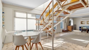 Una sala de estar moderna con una mesa redonda blanca, cuatro sillas grises, una escalera de madera y un gran ventanal con persianas.