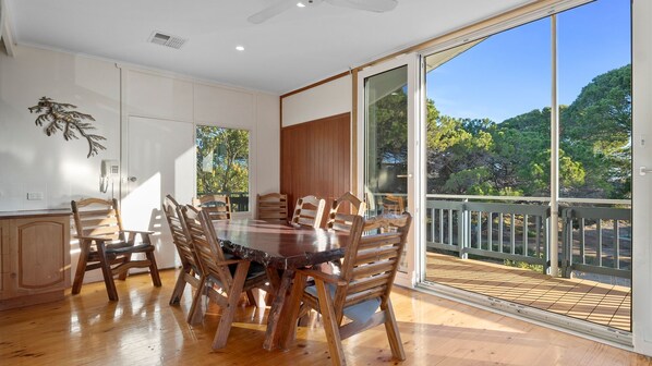 Un coin repas avec une table et des chaises en bois, de grandes fenêtres et une vue sur les arbres et une terrasse.